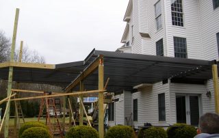 black pergola being constructed for a white home with bushes. ladders are underneath the pergola